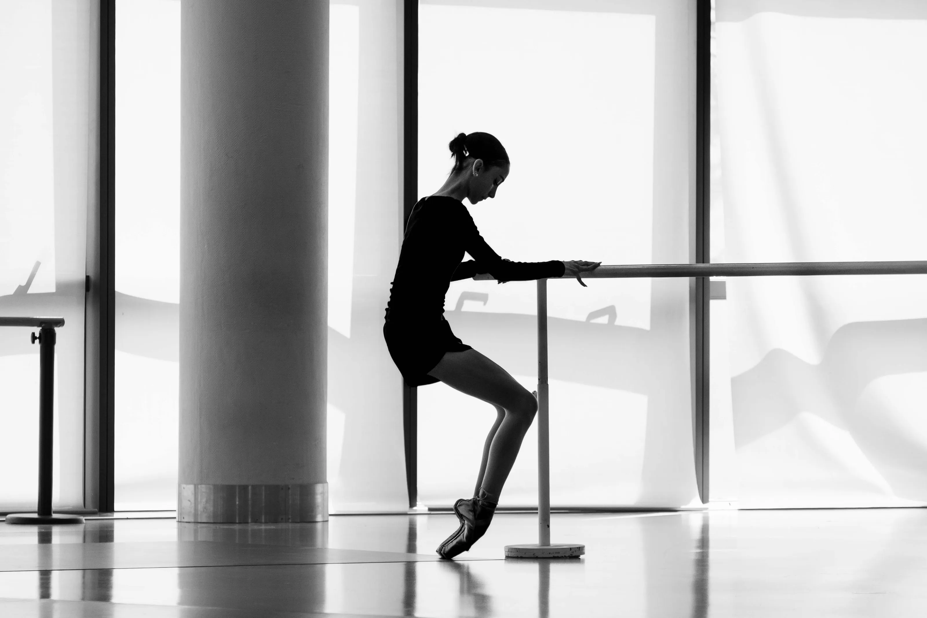 Una ballerina in controluce alla sbarra, in punta, davanti a grandi finestre