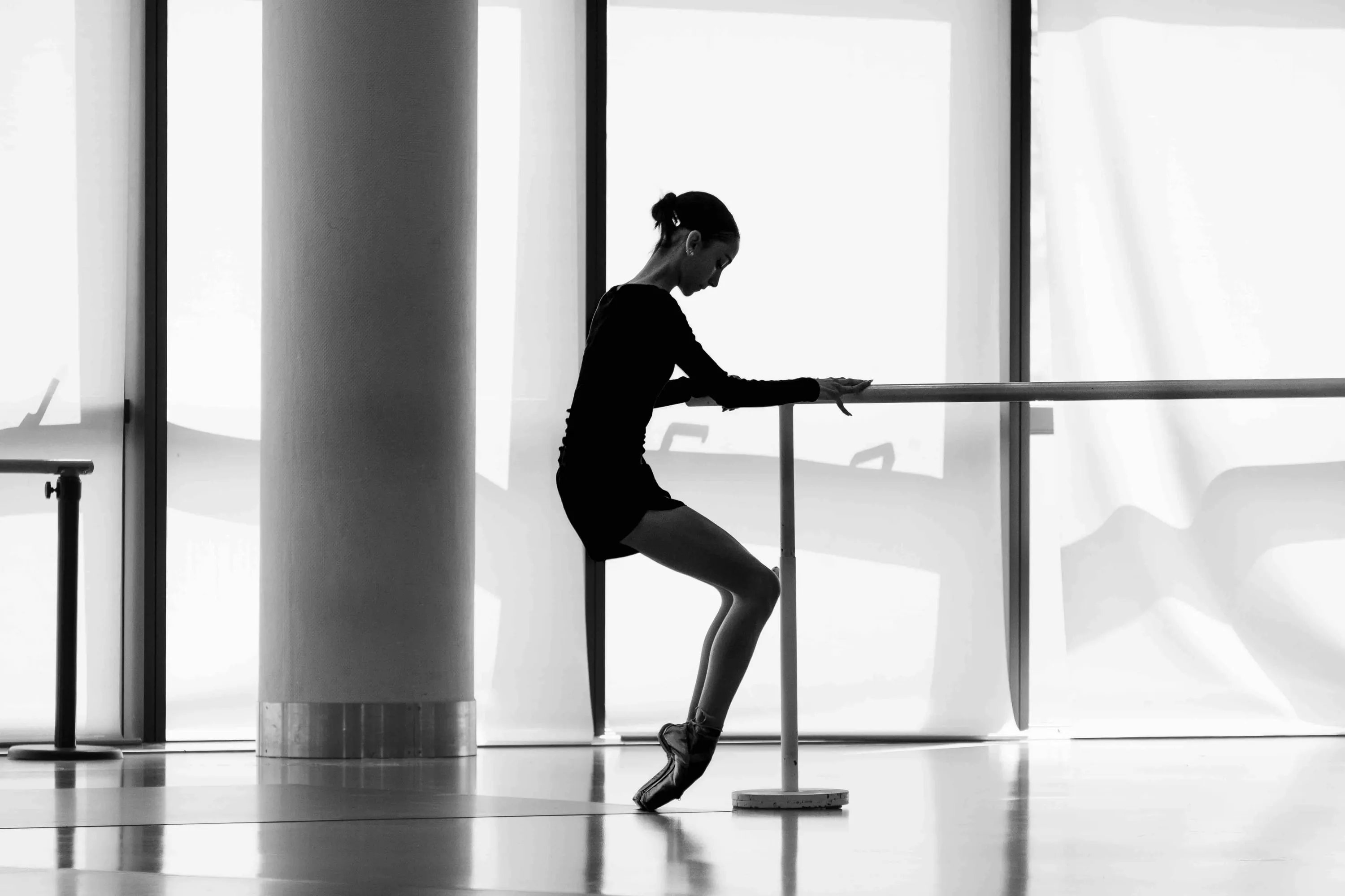 Una ballerina in controluce alla sbarra, in punta, davanti a grandi finestre
