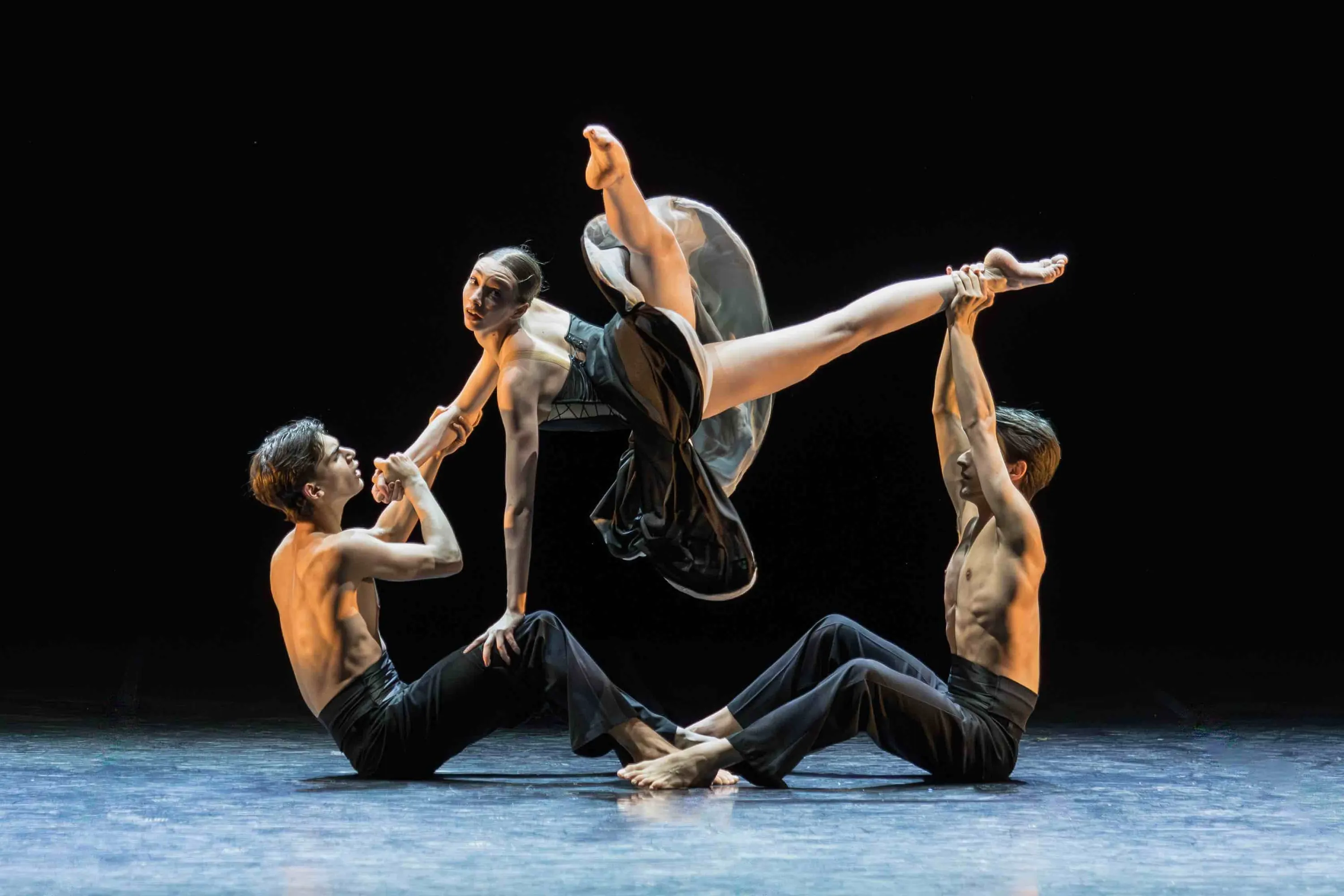 Tre danzatori sollevano una ballerina in aria, su fondo nero